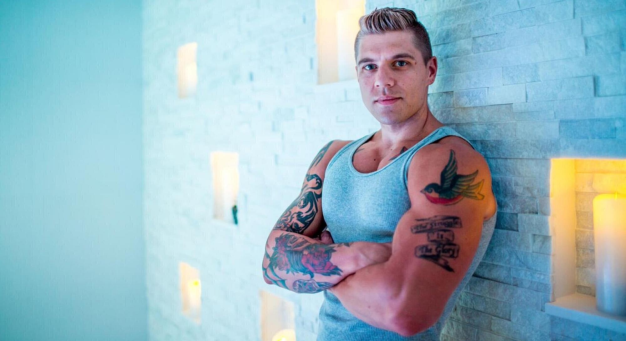 Man with tattoos posing against a light wall.